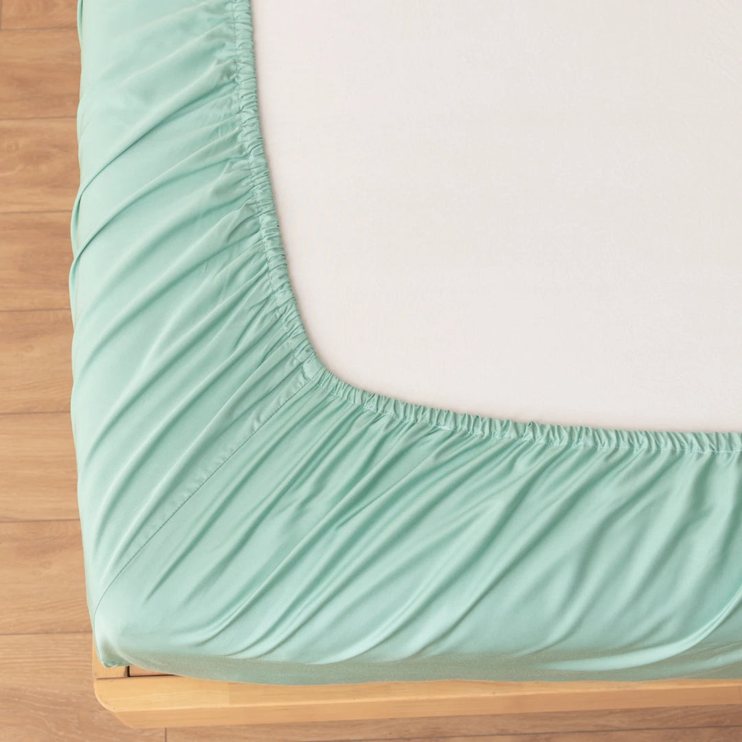 A close-up shows the Linenly Bamboo Fitted Sheet in Summer Green, its elastic edge snugly wrapped around a mattress corner, atop a light wood bed frame with a wooden floor.