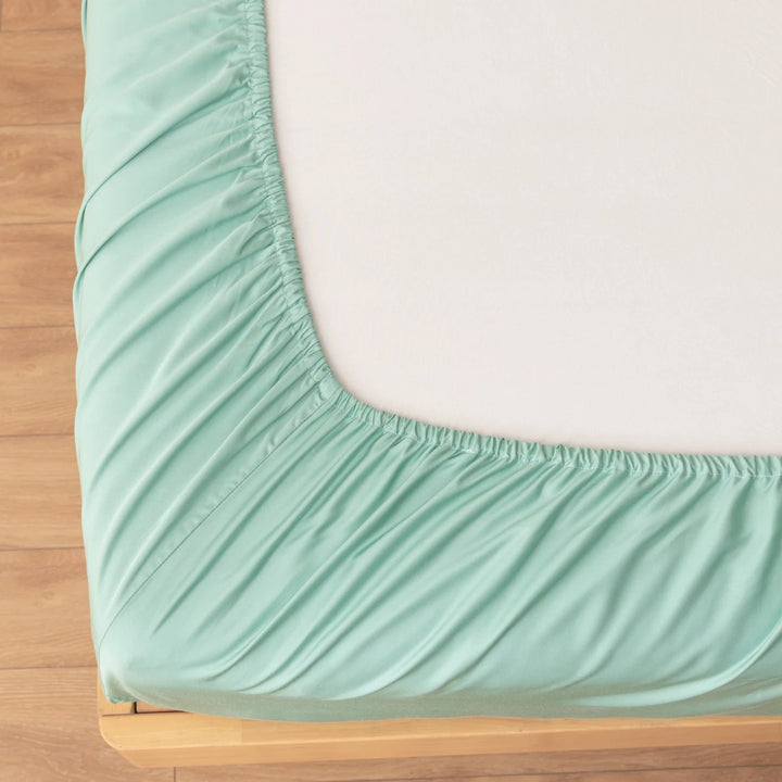 A close-up shows the Linenly Bamboo Fitted Sheet in Summer Green, its elastic edge snugly wrapped around a mattress corner, atop a light wood bed frame with a wooden floor.