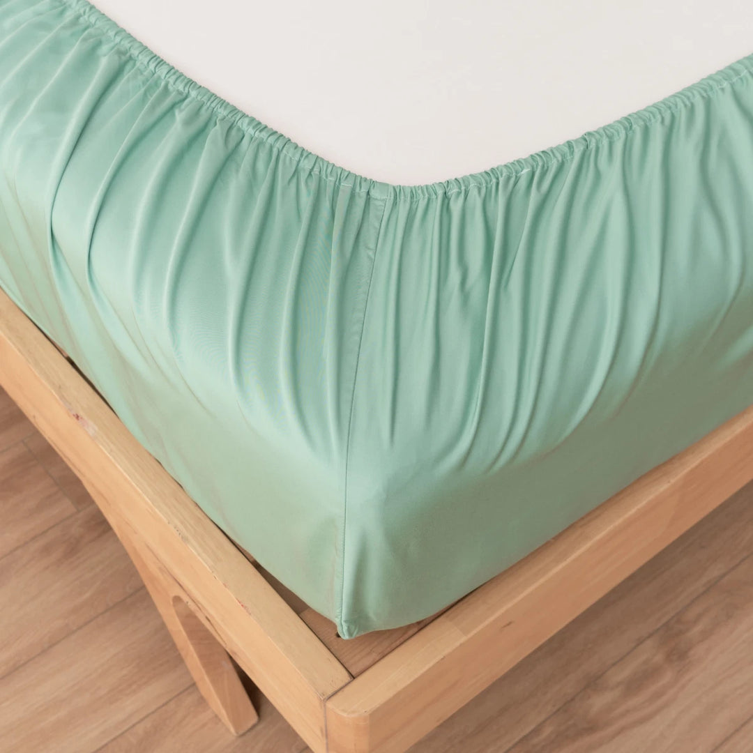 Close-up of Linenly’s Bamboo Fitted Sheet - Summer Green, eco-friendly with deep pockets, snugly covering a mattress corner on a wooden bed frame and hardwood floor.