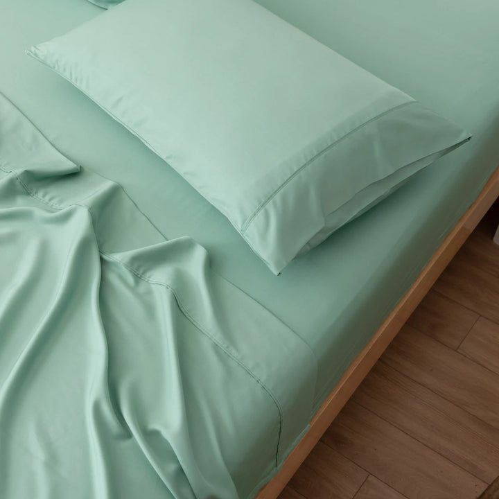 A neatly made bed features Linenly’s Bamboo Fitted Sheet in Summer Green, with matching pillowcase. The deep pockets keep the eco-friendly sheets smooth despite some rumples. The light wood bed frame and floor complement the fresh look.