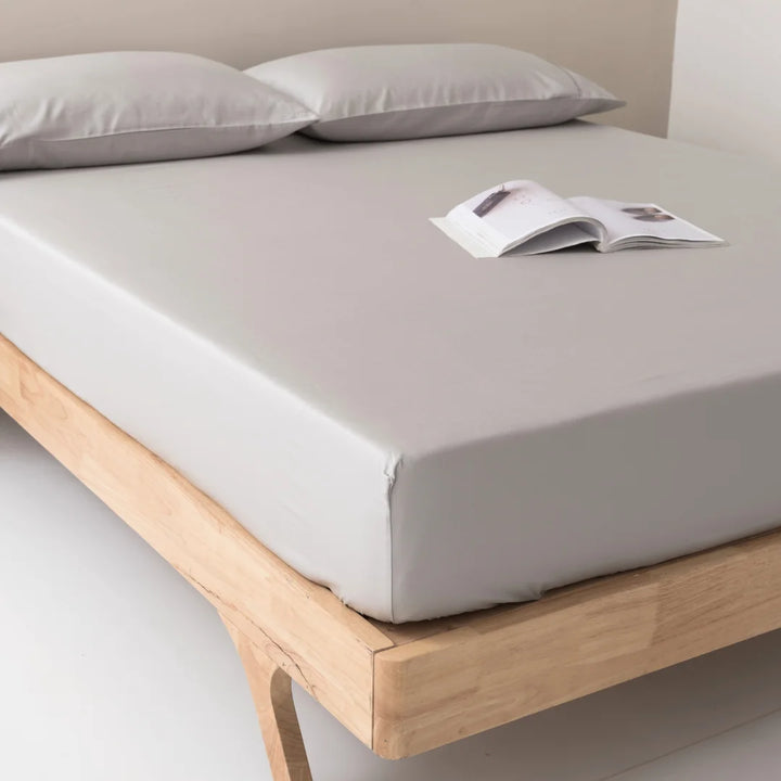 A modern minimalist bedroom featuring a neatly made bed with Linenly's Silver Bamboo Fitted Sheet, two pillows, and an open book resting on the bed, suggesting a quiet, cozy reading spot.