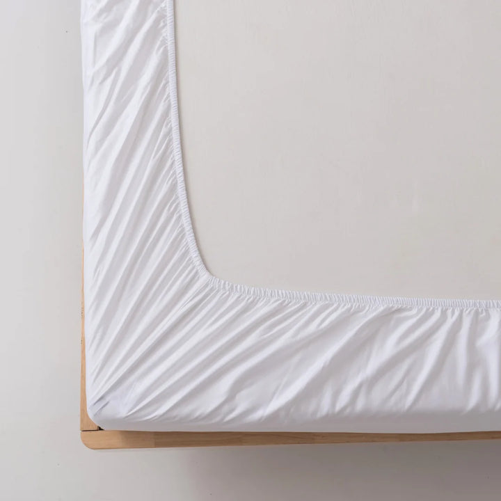 Close-up view of a Linenly Bamboo Fitted Sheet - White on a mattress, showcasing the corner with deep sides on a light wooden bed frame against a neutral background.