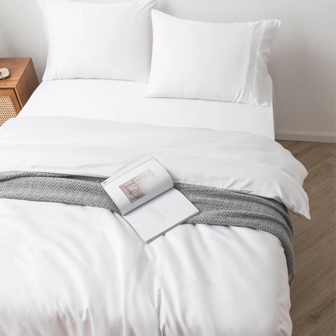 A neatly made bed with Linenly's crisp white bamboo quilt cover, accented by a gray knitted throw blanket at the foot, and an open book resting on the blanket, inviting a peaceful reading session.