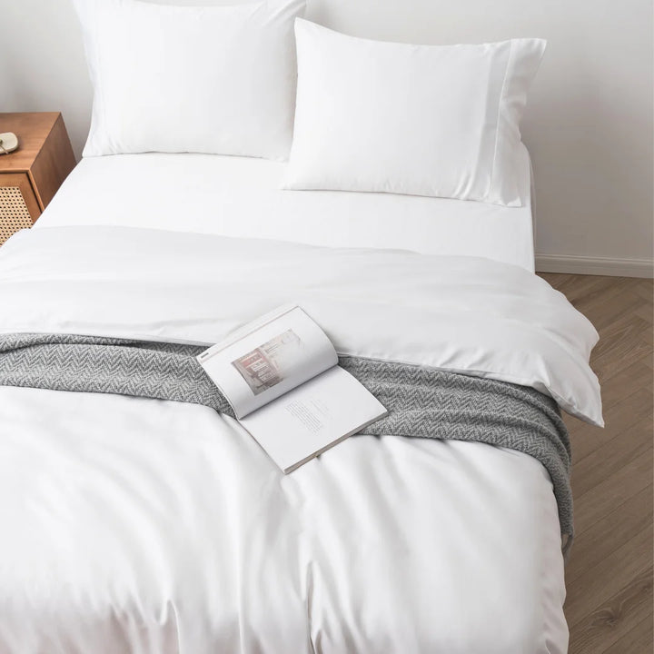 A neatly made bed with Linenly's crisp white bamboo quilt cover, accented by a gray knitted throw blanket at the foot, and an open book resting on the blanket, inviting a peaceful reading session.