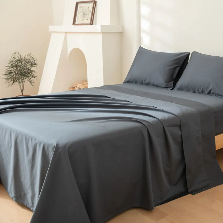 A neatly made bed with smooth, Linenly Luxe Sateen Flat Sheet in Charcoal in a tranquil bedroom setting, featuring a minimalist fireplace and a small potted plant adding a touch of greenery.