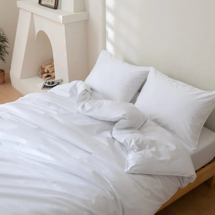 A neatly made bed with crisp white bedding in a bright, minimalist bedroom, featuring an inviting arrangement of fluffy pillows encased in Linenly Luxe Sateen Pillowcase Set - White and a clean, tranquil ambiance.