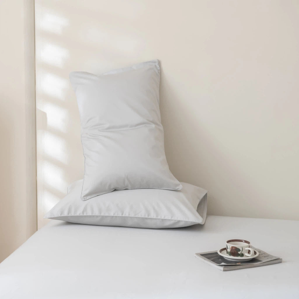 A cozy, minimalist bedroom features a white bed with two neatly stacked pillows against a light wall. Soft sunlight filters through blinds, casting shadows. A small tray holds a cup and notebook on the bed.