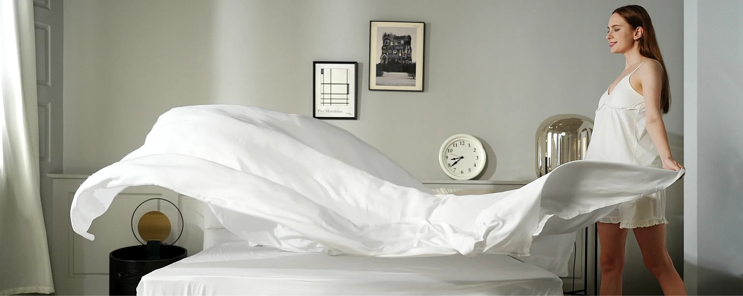 A woman in a white dress makes a bed, gracefully tossing a white sheet over it. The room is minimalistic with framed art, a round table with a lamp, and a clock on the wall.