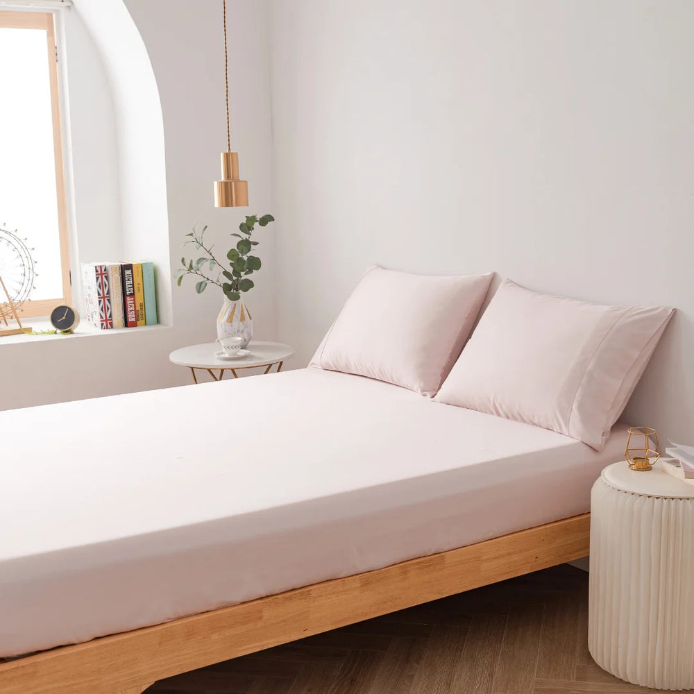 A serene and minimalist bedroom design featuring a low wooden bed frame with a white mattress, soft pink pillows, complemented by a white round bedside table with books, a plant, and a hanging pendant light from Linenly's Bamboo Fitted Sheet in Blush.