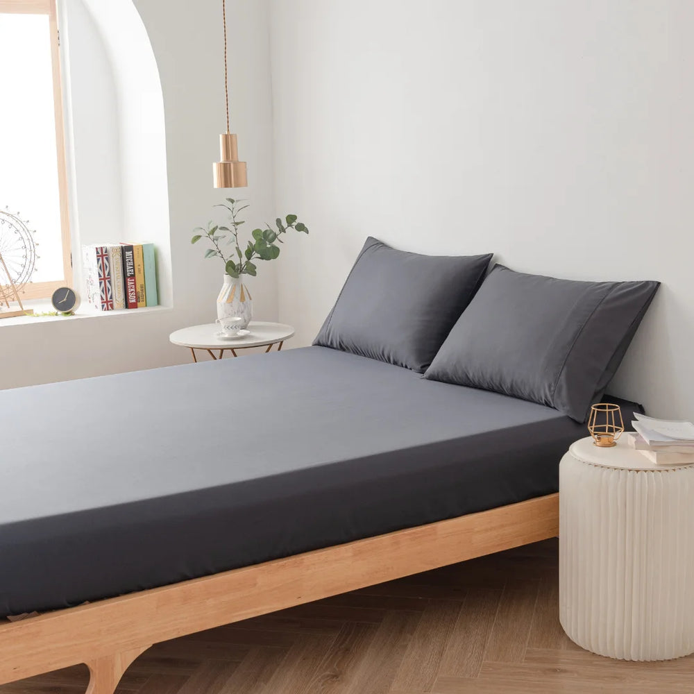 Minimalist bedroom interior with a modern wooden bed, Linenly charcoal bamboo fitted sheet, and a small bedside table featuring a vase with greenery, books, and a clock under an arched window.