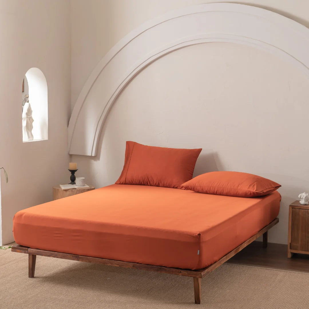 A minimalist bedroom highlights a wooden bed frame with Linenly's Bamboo Fitted Sheet in Copper Sunset, providing luxury and comfort. An orange pillow pops against a white arched wall accent, while decorations sit in a window niche, and the floor is covered by a beige carpet.