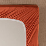 The Bamboo Fitted Sheet - Copper Sunset by Linenly snugly wraps around a beige mattress in a soothing orange hue, creating smooth lines and blending seamlessly with the light brown carpeted floor beneath.