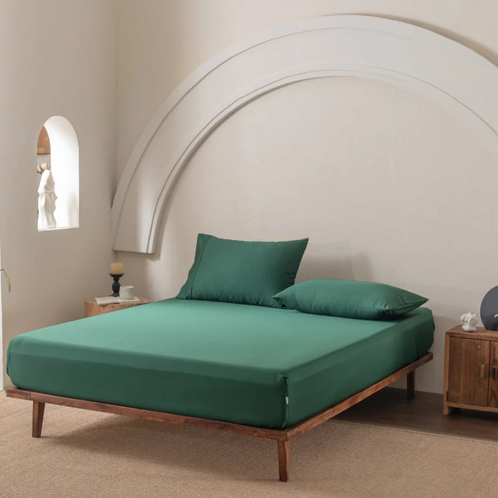 A minimalist bedroom showcases a wooden bed with a green bedspread, enhanced by Linenly's Bamboo Fitted Sheet Set in Evergreen. Set against a cream wall with an arched feature, matching pillows and luxury bedding add comfort. A wooden nightstand and woven rug complete the serene look.