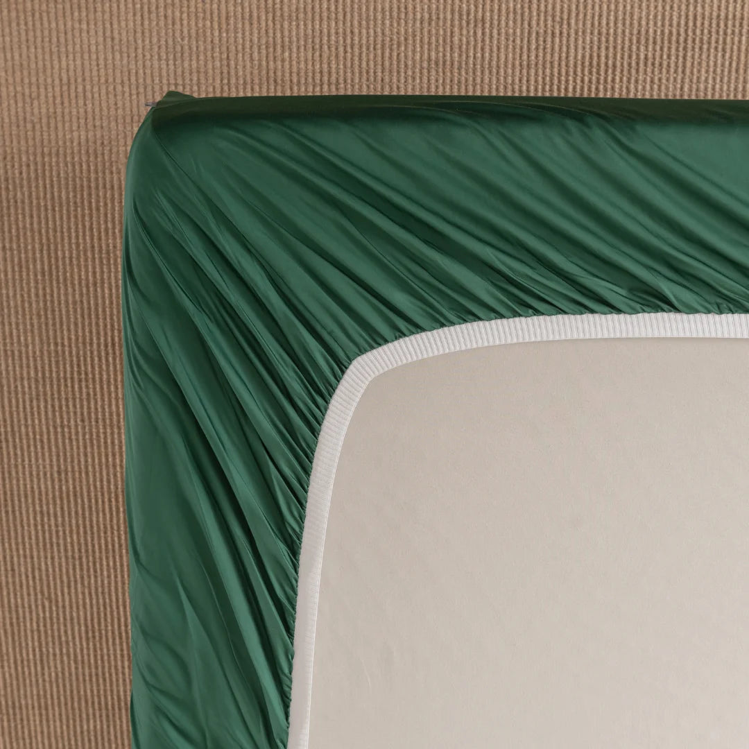 A top view shows a corner of a mattress draped with the Bamboo Fitted Sheet - Evergreen by Linenly, partially revealing its soft, light-colored top and white border. The mattress sits on a textured beige surface, creating an elegant contrast.