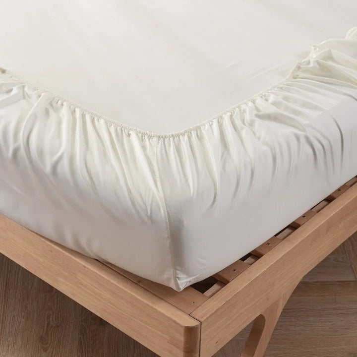 A close-up view of neatly fitted Linenly Ivory Bamboo Fitted Sheets on a wooden bed frame, showcasing the elastic edges securing the sheet to the mattress—an eco-friendly choice.