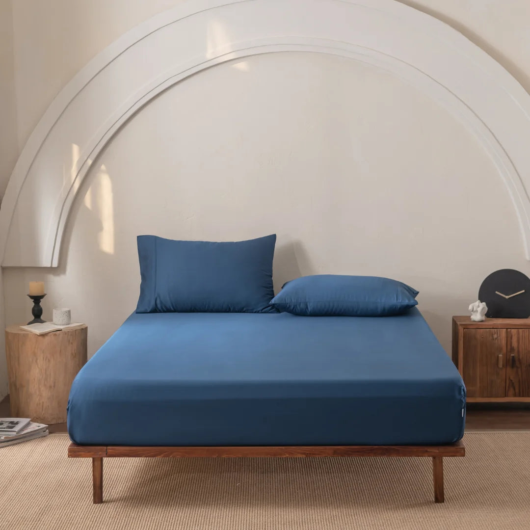 A minimalist bedroom showcases a wooden bed frame with Linenly Bamboo Fitted Sheet Set in Midnight and two matching pillows, flanked by wooden side tables with a candle, clock, and book. An arched wall design enhances the decor.