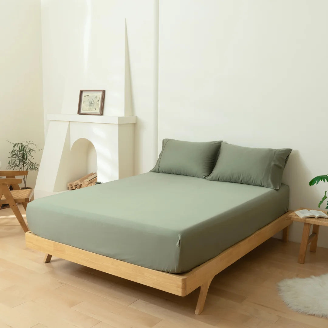 A minimalist bedroom with a low-profile wooden bed, dressed in Linenly Bamboo Fitted Sheet in Moss, against a soft white wall with a decorative fireplace and a framed picture on the mantel, complemented by greenery.