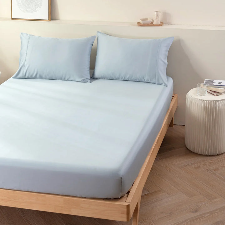 A minimalistic bedroom with a wooden bed frame featuring neatly arranged Linenly Bamboo Fitted Sheet Set in pale blue and matching pillows. Beside the bed is a small, white round bedside table adorned with decorative items and a cup. The room exudes an eco-friendly, light, and airy ambiance with a cooling feel.