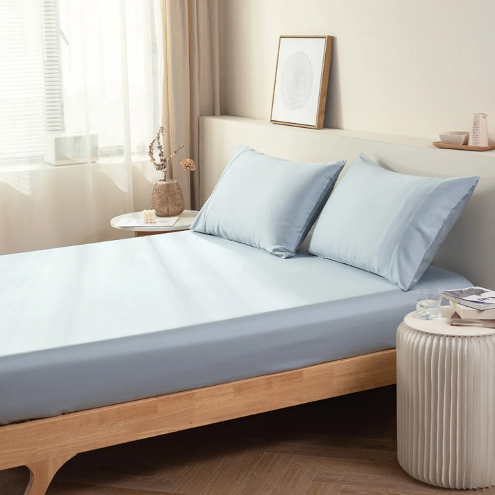 A neatly made bed with a Linenly Bamboo Fitted Sheet Set in Pale Blue and pillows stands in a minimalistic bedroom. A round side table with a vase of dried flowers and a book is placed next to the bed. Sunlight filters through sheer curtains on the window, creating an eco-friendly, serene atmosphere with a cooling feel.