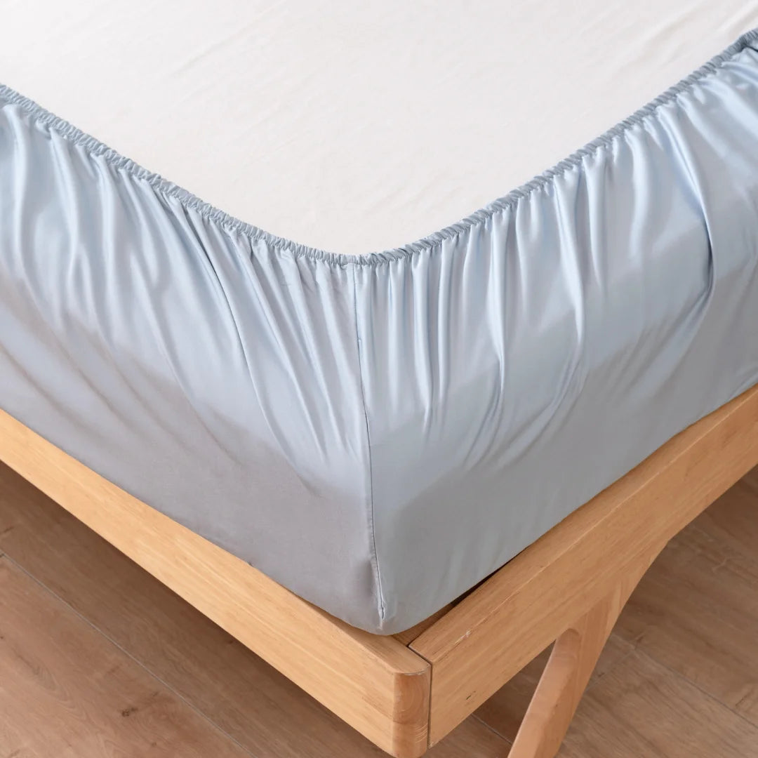 A close-up of a bed with a wooden frame. The bed is adorned with the Bamboo Fitted Sheet Set in Pale Blue from Linenly, snugly wrapped around the mattress, offering an eco-friendly and cooling feel. The room has a wooden floor.