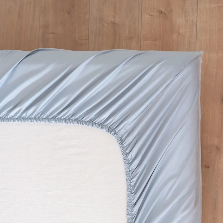 A close-up of a bed features Linenly's Bamboo Fitted Sheet Set in Pale Blue, partially covering a white mattress. The corner of the eco-friendly sheet is visibly stretched over the mattress corner, creating a cool feel against the wooden floor background.