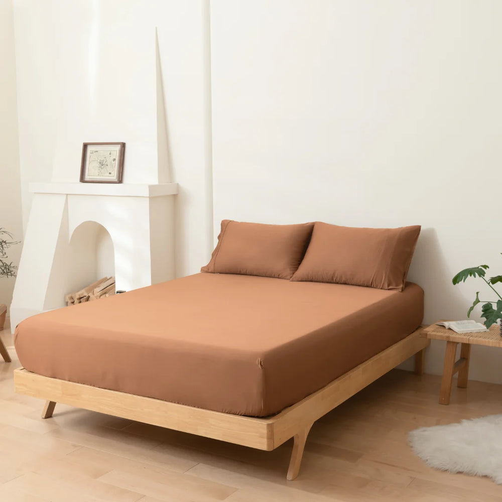 Minimalist bedroom with a low-profile wooden bed frame and matching Linenly Terracotta Bamboo Fitted Sheet, complemented by a white decorative fireplace, framed picture, and a touch of greenery.