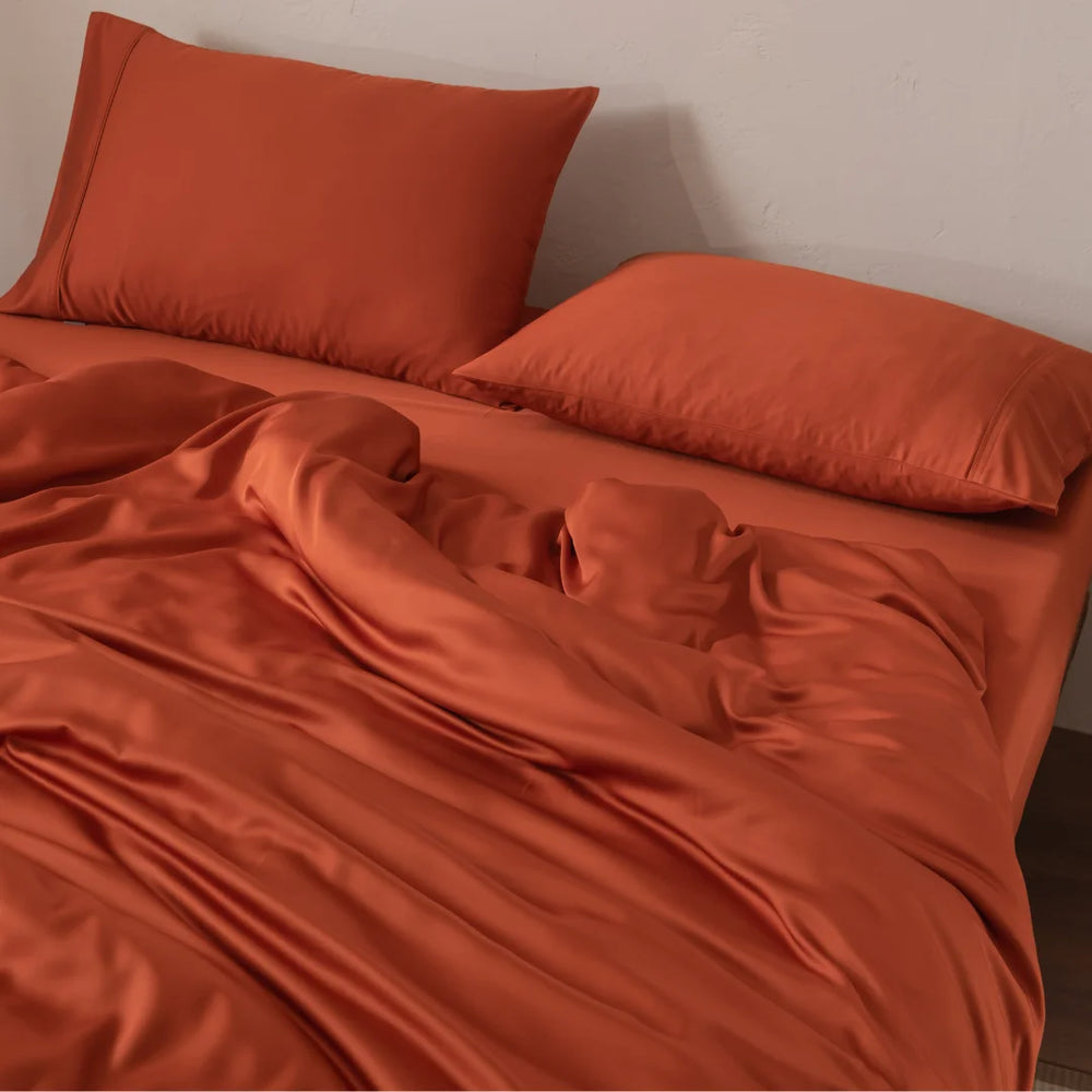 A bed featuring Linenly's Bamboo Pillowcase Set in Copper Sunset, with orange sheets and a comforter, is slightly disheveled against a light wall. The vibrant bedding creates a warm, inviting atmosphere enhanced by the silky-smooth feel of organic bamboo fabric.