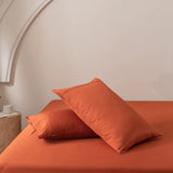 Two pillows in Linenly's Bamboo Pillowcase Set - Copper Sunset rest on an orange bedspread against a light beige wall. A wooden bedside table with a small plant and candle is partially visible on the left, completing the minimalistic and cozy setting.