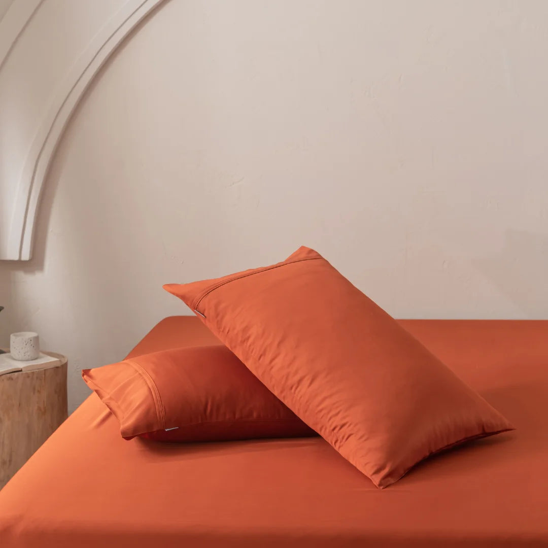 Two pillows in Linenly's Bamboo Pillowcase Set - Copper Sunset rest on an orange bedspread against a light beige wall. A wooden bedside table with a small plant and candle is partially visible on the left, completing the minimalistic and cozy setting.