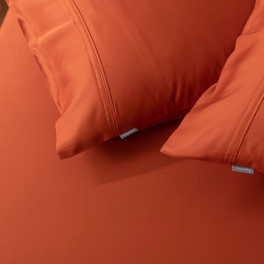 Two Bamboo Pillowcase Set - Copper Sunset from Linenly with visible seams rest on an orange bed sheet, creating a cozy monochromatic look, while the satin weave and small edge tags add an elegant touch.