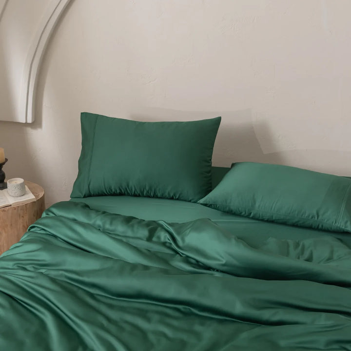 A neatly made bed showcases smooth, dark green satin weave bedding and Linenly's Bamboo Pillowcase Set - Evergreen. Two large pillows rest invitingly, while a wooden bedside table holds a small white candle in a glass jar. The wall's curved design adds to the cozy atmosphere.