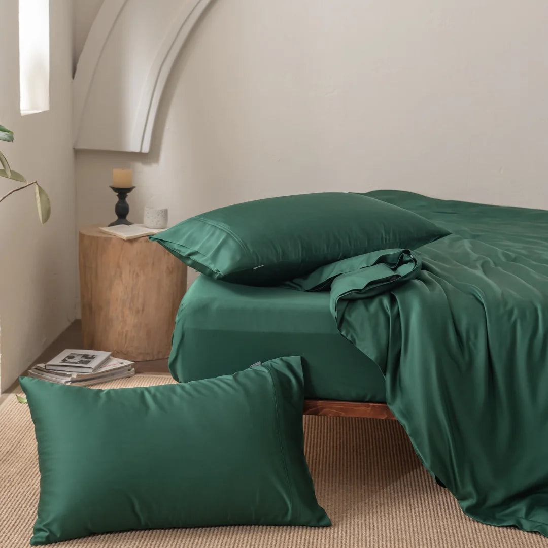 A bedroom showcases a bed adorned with satin weave dark green sheets and Linenly Bamboo Pillowcase Set - Evergreen. A matching pillow lies on the beige carpet. A wooden nightstand holds a candle and books, while an organic bamboo potted plant is visible in the corner.