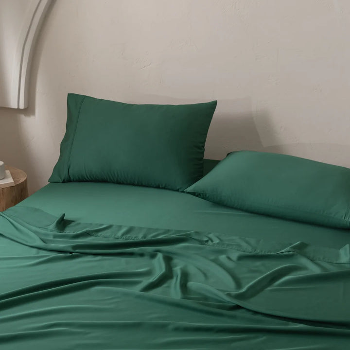 A neatly made bed showcases deep green satin weave bedding with two Linenly Bamboo Pillowcase Set - Evergreen. A small wooden nightstand beside it holds a white item, and the light-colored wall features a subtle arch design to elegantly enhance the room.