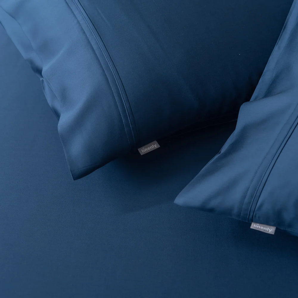 Deep blue pillows wrapped in Linenly's Bamboo Pillowcase Set - Midnight rest on a matching blue sheet, showcasing the luxurious satin weave fabric. The small tag on each pillow adds a touch of simplicity and comfort to the image.