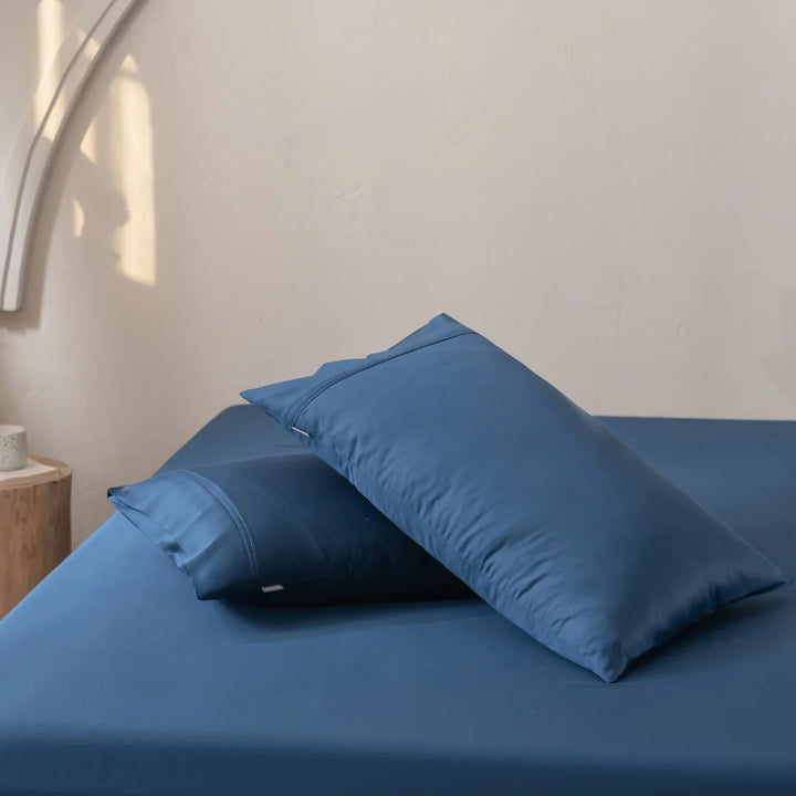 Two blue satin weave pillows rest on a bed with Linenly's Bamboo Pillowcase Set in Midnight and matching blue sheets. Positioned near a wooden side table, the off-white wall backdrop enhances a calm, minimalist vibe.