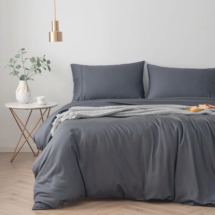 Modern bedroom with a neatly made bed featuring an eco-friendly Linenly bamboo quilt cover in charcoal, complemented by a small side table with a plant and a cozy throw blanket, creating a tranquil and minimalist atmosphere.