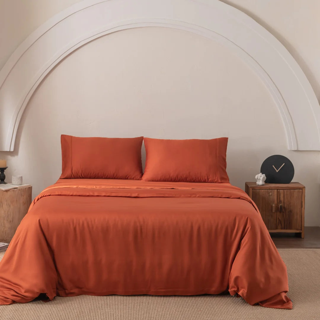 A neatly made bed with a Linenly Bamboo Quilt Cover in Copper Sunset and two matching pillows is centered in a room under a large white arch. Flanking the bed are two wooden nightstands, one featuring a round black clock, all resting on a beige carpeted floor.