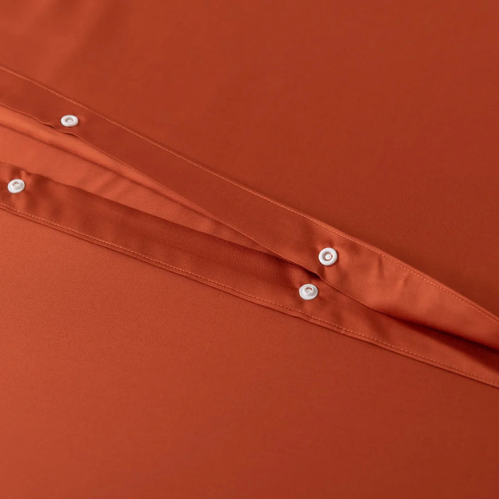 Close-up of the Bamboo Quilt Cover in Copper Sunset by Linenly, showcasing an orange fabric with a folded seam and small white round buttons. The silky texture adds a premium feel, while the neat stitching offers a clean, tailored look.