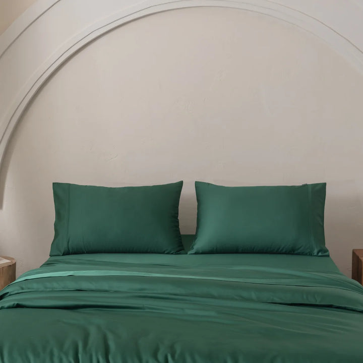 A neatly made bed with Linenly's Bamboo Quilt Cover - Evergreen and green sheets lies under an arched white wall. Wooden nightstands on each side enhance the serene, minimalist space crafted from premium bamboo materials.