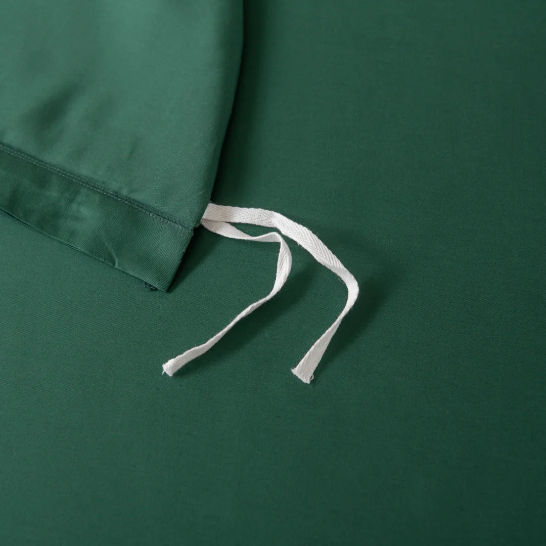 A close-up of Linenly's Bamboo Quilt Cover Set in Evergreen shows its silky texture with a drawstring edge on a flat surface. Two white strings tied against the solid green background evoke a restful sleep experience.