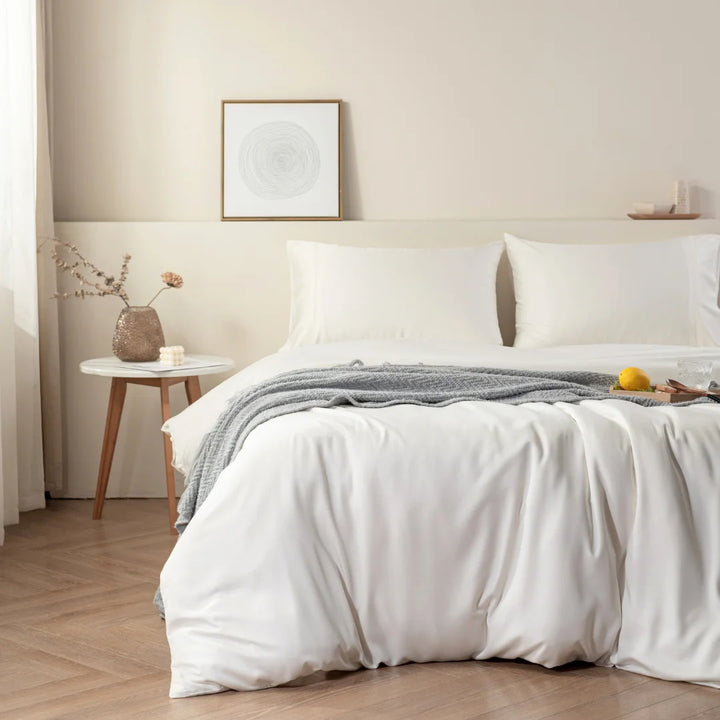 A cozy and neatly made bed with a Linenly Ivory Bamboo Quilt Cover and gray bedding in a minimalist bedroom, featuring a small wooden side table with a vase and flowers, and a framed abstract artwork on the wall.