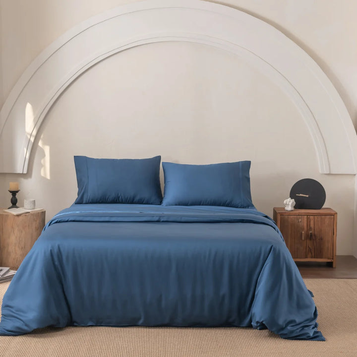 The bedroom showcases the Linenly Bamboo Quilt Cover Set - Midnight, known for its luxurious silky feel. Two pillows complement the look, while wooden nightstands hold decor items. A curved white wall accent and beige carpet flooring enhance the room's calm and minimalist atmosphere.