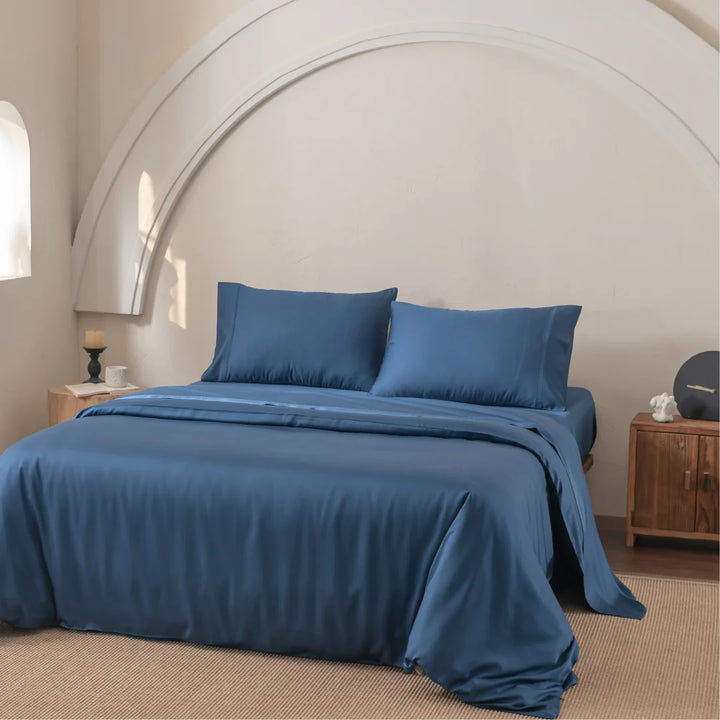 A Linenly Bamboo Quilt Cover in Midnight adorns the centered bed, exuding supreme softness. A white, curved architectural wall feature complements its silky texture. A wooden side table with a candle is on one side and a cabinet on the other, atop a carpeted floor.