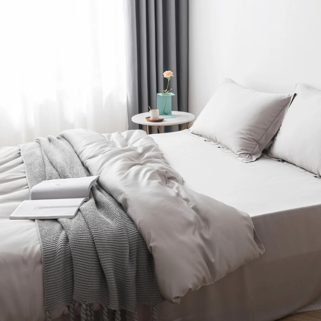 A cozy and neatly arranged bedroom with a luxurious Linenly silver bamboo quilt cover, an inviting open laptop waiting on the bed, and a small bedside table sporting a single pink flower in a vase, creating a serene atmosphere.