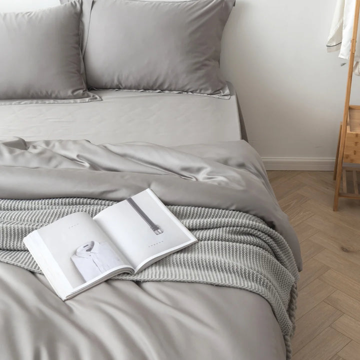 A serene bedroom corner with a neatly made bed draped with a Linenly Stone Grey bamboo quilt cover and an open book laid invitingly on it, beckoning for a lazy afternoon read.