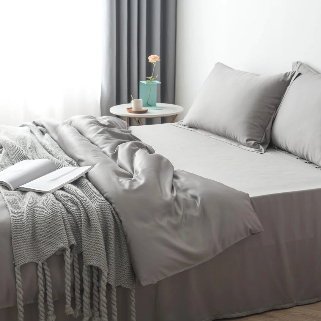 A serene bedroom corner with an unmade bed covered in Linenly's stone grey bamboo quilt cover, featuring a knit throw blanket casually draped over, a single rose in a vase on a bedside table, and an open.