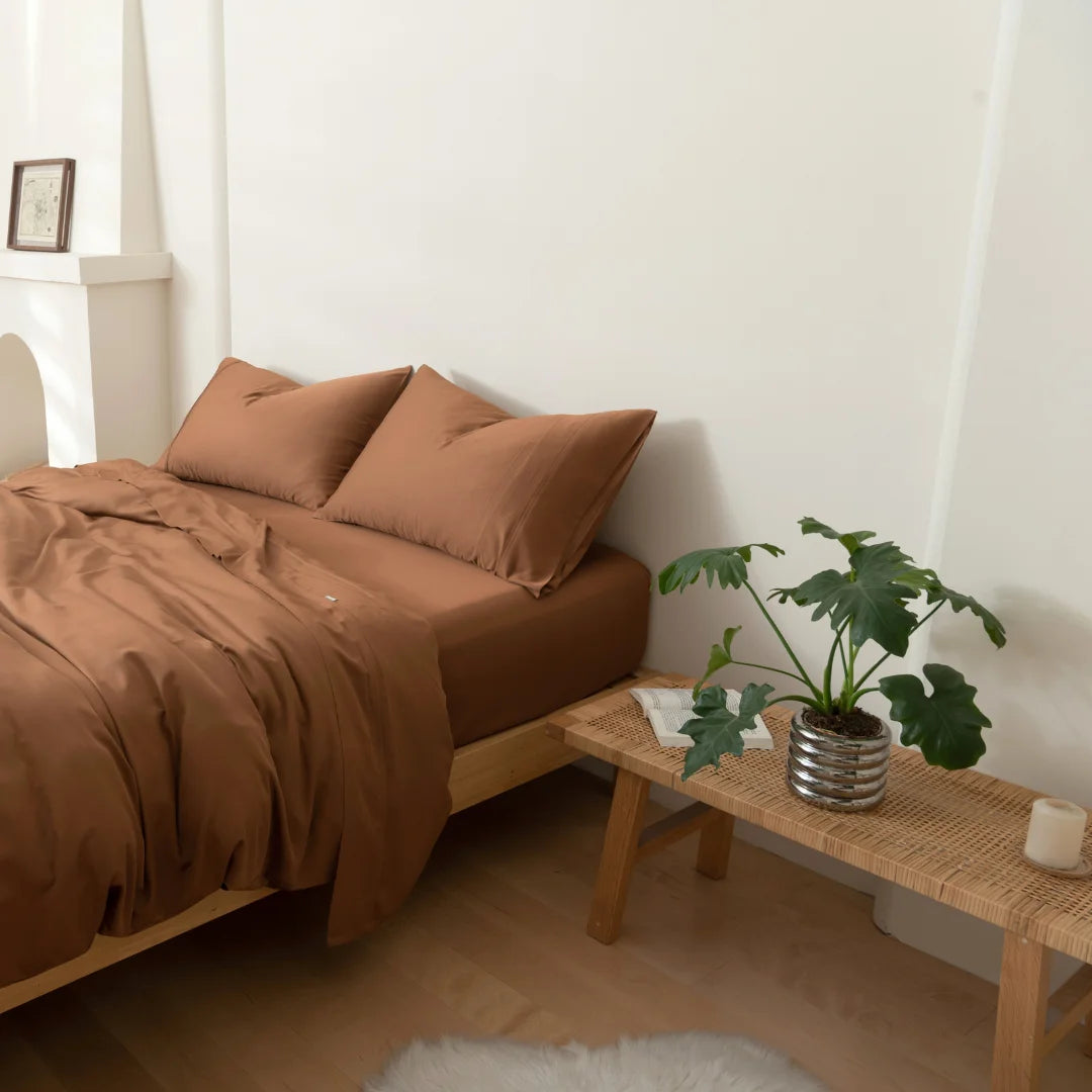 A neatly made bed with the Linenly Bamboo Quilt Cover Set in Terracotta sits beside a woven bench holding a potted plant and candle. Light walls and floors enhance the calm, minimalist space with eco-friendly bedding from Linenly.
