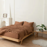 A minimalist bedroom with a wooden bed frame and brown bedding, featuring the Linenly Bamboo Quilt Cover Set in Terracotta, two pillows, a small plant on a wooden bench, a white rug, and a simple white fireplace with a framed picture.