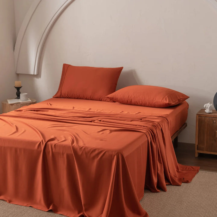 A minimally decorated room features a bed with Linenly's Bamboo Sheet Set in Copper Sunset and two orange pillows. The luxuriously smooth sheets drape elegantly, complemented by a wooden nightstand holding a candle and decor beneath a white wall featuring a curved arch.