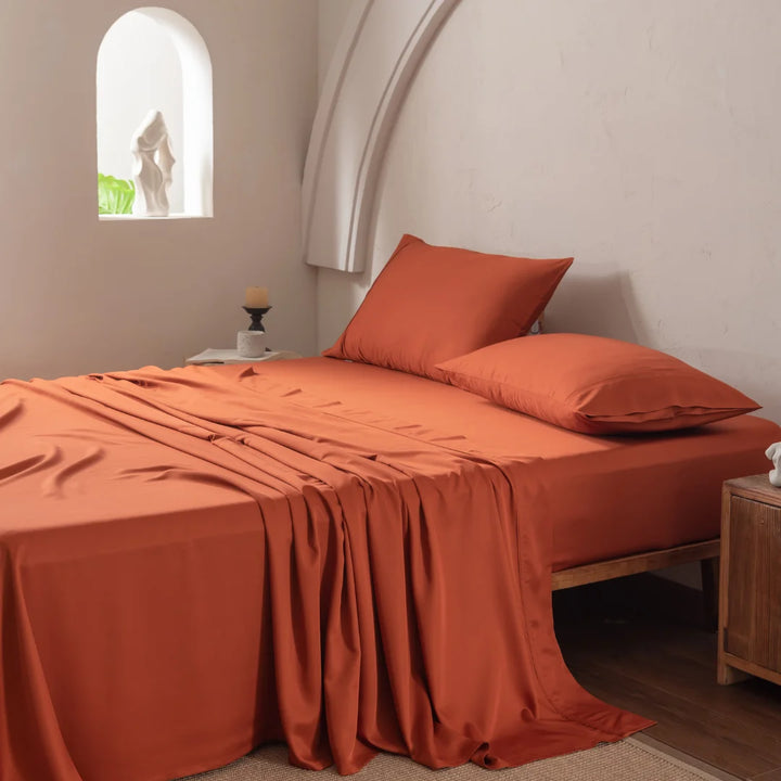 A cozy bed adorned with Linenly's Bamboo Sheet Set in Copper Sunset and pillows stands against a white wall, featuring an arched niche with a sculpture. A wooden nightstand is topped with a candle, and soft natural light fills the room.